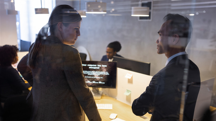 A man and a woman stand in a professional setting, looking at one another as they try to solve a technical problem